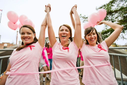 Three young women dressed in pink run in marathon to fight breast cancer. They joyfully cross the finish line at the end of a bridge with pink balloons